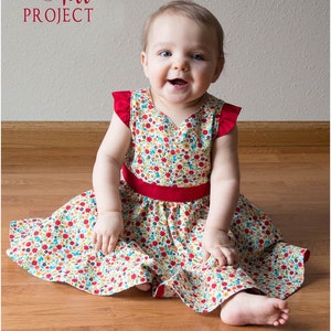 May include: A baby girl wearing a white dress with a red floral pattern and a red sash. The dress has short sleeves with red ruffles. The baby is sitting on a wooden floor. The text "Sew & Tell Project" is in the top left corner of the image. The text "Peek-a-Boo Pattern Shop" is in the bottom left corner of the image.