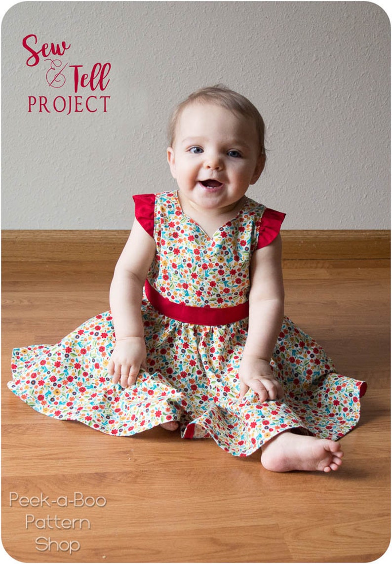 May include: A baby girl wearing a white dress with a red floral pattern and a red sash. The dress has short sleeves with red ruffles. The baby is sitting on a wooden floor. The text "Sew & Tell Project" is in the top left corner of the image. The text "Peek-a-Boo Pattern Shop" is in the bottom left corner of the image.