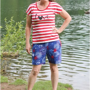 Pu&ograve; includere: Una donna indossa una maglietta a righe rosse e bianche con la scritta "Love" e una silhouette di Topolino sul davanti. Indossa anche pantaloncini blu e bianchi con un motivo a fuochi d'artificio. &Egrave; in piedi davanti a un lago.