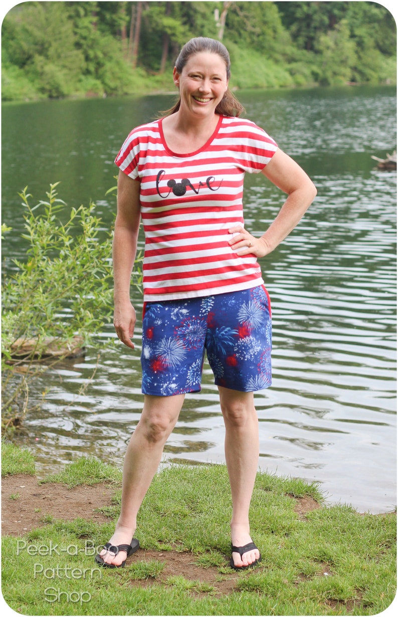 Pu&ograve; includere: Una donna indossa una maglietta a righe rosse e bianche con la scritta "Love" e una silhouette di Topolino sul davanti. Indossa anche pantaloncini blu e bianchi con un motivo a fuochi d'artificio. &Egrave; in piedi davanti a un lago.