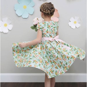 May include: A young girl wearing a light blue floral dress with a pink sash and white sandals. The dress has a full skirt and short sleeves. The girl is standing in front of a wall decorated with paper flowers.