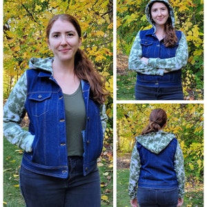 May include: A blue denim hooded jacket with green and white camouflage sleeves. The jacket has a button-up front and pockets. The person in the image is wearing a green shirt and black jeans. The background features fall foliage.