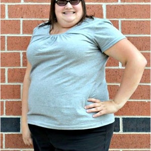 May include: A woman wearing a gray t-shirt with a scoop neck and black pants. She is standing in front of a brick wall and is wearing sunglasses.