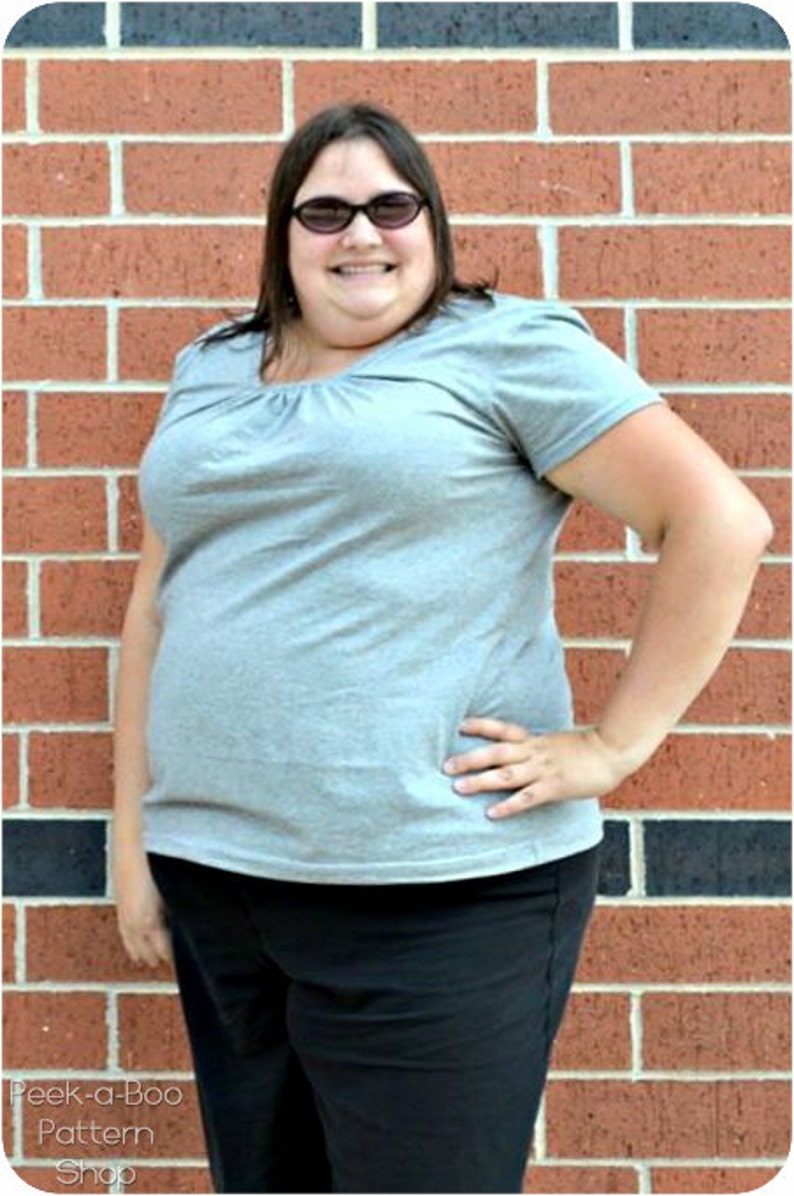 May include: A woman wearing a gray t-shirt with a scoop neck and black pants. She is standing in front of a brick wall and is wearing sunglasses.