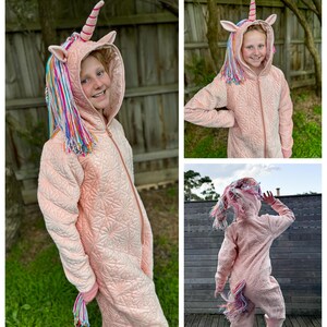 May include: A pink unicorn onesie with a rainbow mane and tail. The onesie has a zipper down the front and a hood with unicorn ears. The child wearing the onesie is smiling and looking at the camera.