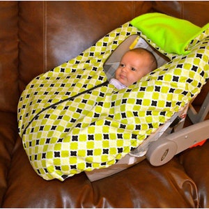 May include: A baby in a green and black patterned car seat cover. The cover has a hood that can be pulled up to protect the baby from the sun. The car seat is on a brown leather couch.