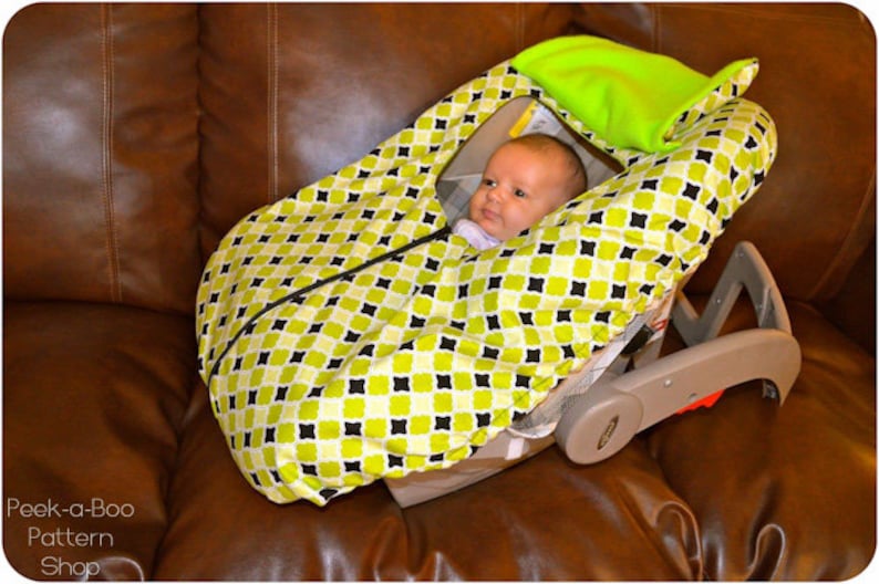 May include: A baby in a green and black patterned car seat cover. The cover has a hood that can be pulled up to protect the baby from the sun. The car seat is on a brown leather couch.