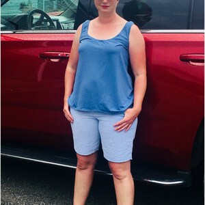 Pu&ograve; includere: Una donna indossa una canotta blu e pantaloncini blu chiaro, in piedi accanto a un'auto rossa. Indossa occhiali da sole bianchi e ha i capelli corti castani.