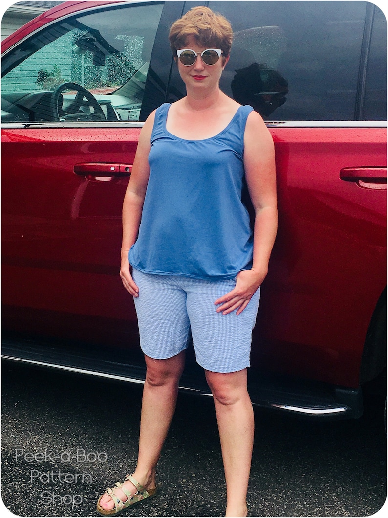 Pu&ograve; includere: Una donna indossa una canotta blu e pantaloncini blu chiaro, in piedi accanto a un'auto rossa. Indossa occhiali da sole bianchi e ha i capelli corti castani.