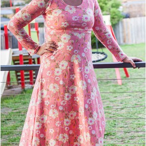 May include: A woman wearing a pink maxi dress with a floral print. The dress has long sleeves and a round neckline. The woman is standing on a wooden deck.