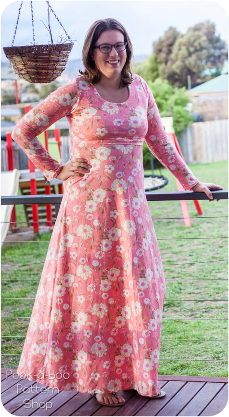 May include: A woman wearing a pink maxi dress with a floral print. The dress has long sleeves and a round neckline. The woman is standing on a wooden deck.