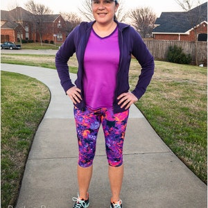 May include: A woman wearing athletic apparel, including a purple zip-up hoodie, a magenta v-neck shirt, and capri leggings with a colorful floral pattern. She is also wearing a visor and running shoes. The text "Peek-a-Boo Pattern Shop" is visible.