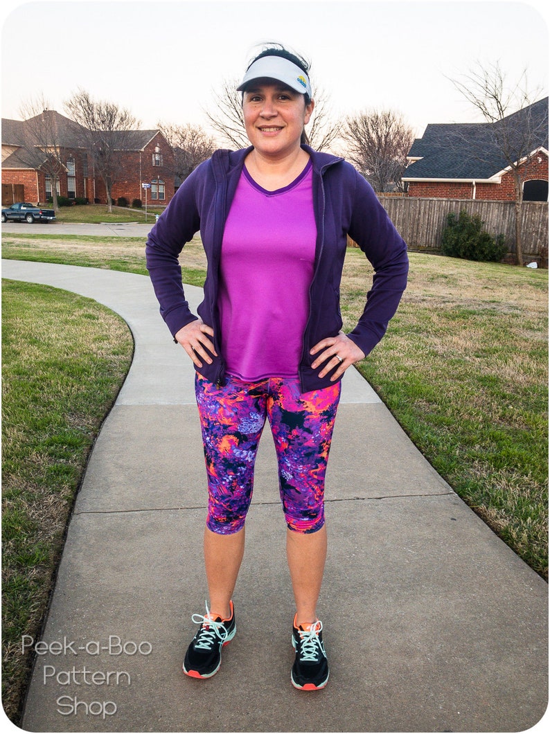 May include: A woman wearing athletic apparel, including a purple zip-up hoodie, a magenta v-neck shirt, and capri leggings with a colorful floral pattern. She is also wearing a visor and running shoes. The text "Peek-a-Boo Pattern Shop" is visible.