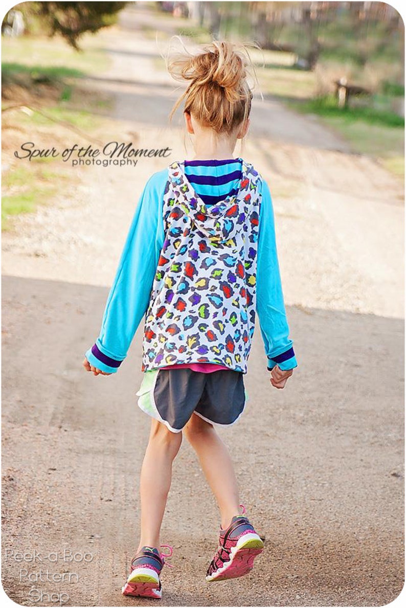 May include: A young person wearing a blue and white hooded sweatshirt with a colorful animal print design. The sweatshirt has a blue and white striped band around the neck. The person is wearing black shorts and pink and black sneakers. The person is walking on a gravel path.