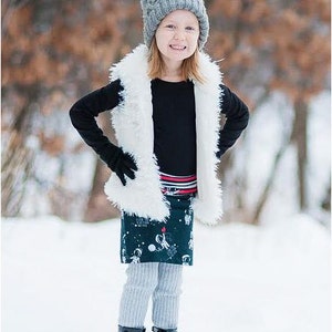 Puede incluir: Una persona joven con un gorro de punto gris, un chaleco de piel sint&eacute;tica blanca, una falda estampada en blanco y negro, medias grises y botas negras. La persona est&aacute; de pie en un entorno exterior nevado.