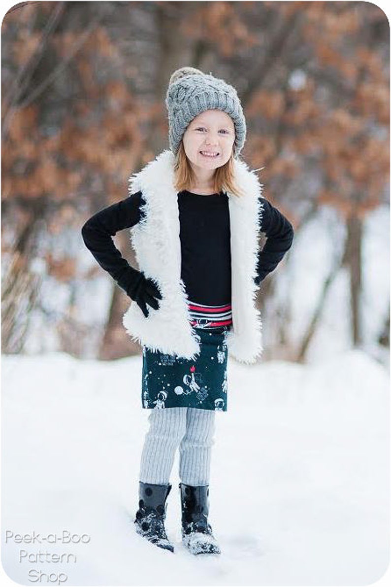 Puede incluir: Una persona joven con un gorro de punto gris, un chaleco de piel sint&eacute;tica blanca, una falda estampada en blanco y negro, medias grises y botas negras. La persona est&aacute; de pie en un entorno exterior nevado.