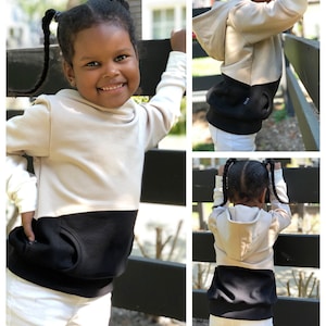 May include: A child wearing a beige and black hooded sweatshirt with a kangaroo pocket. The sweatshirt has a contrasting black bottom and a beige hood.