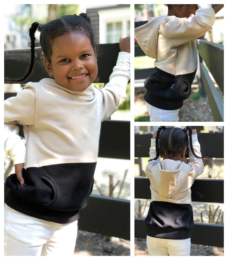 May include: A child wearing a beige and black hooded sweatshirt with a kangaroo pocket. The sweatshirt has a contrasting black bottom and a beige hood.