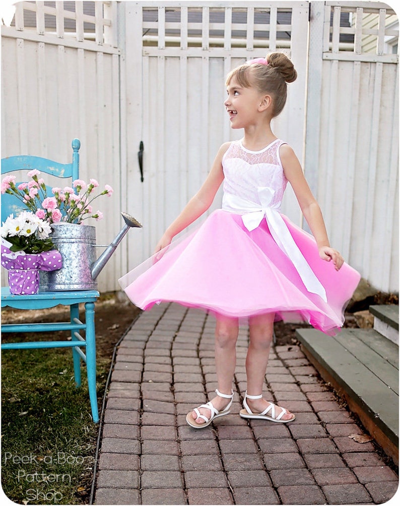 May include: A young girl wearing a pink and white dress with a white sash and a white bow. She is standing on a brick path in front of a white fence. She is smiling and looking to the right.