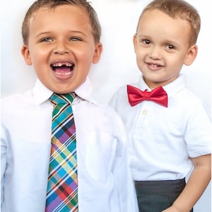 May include: Two young boys wearing white shirts and dress pants. One boy is wearing a red bow tie and the other is wearing a colorful plaid tie. They are both smiling.