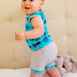 Puede incluir: Un ni&ntilde;o peque&ntilde;o con una camiseta sin mangas turquesa con estampado de telara&ntilde;a y ara&ntilde;a y pantalones cortos grises. El ni&ntilde;o est&aacute; de pie en una cama blanca con un animal de peluche cerca.