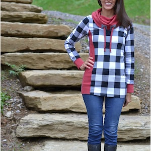 May include: A woman wearing a black and white plaid shirt with a red cowl neck and long sleeves. She is wearing blue jeans and black boots. She is standing in front of a stone staircase.