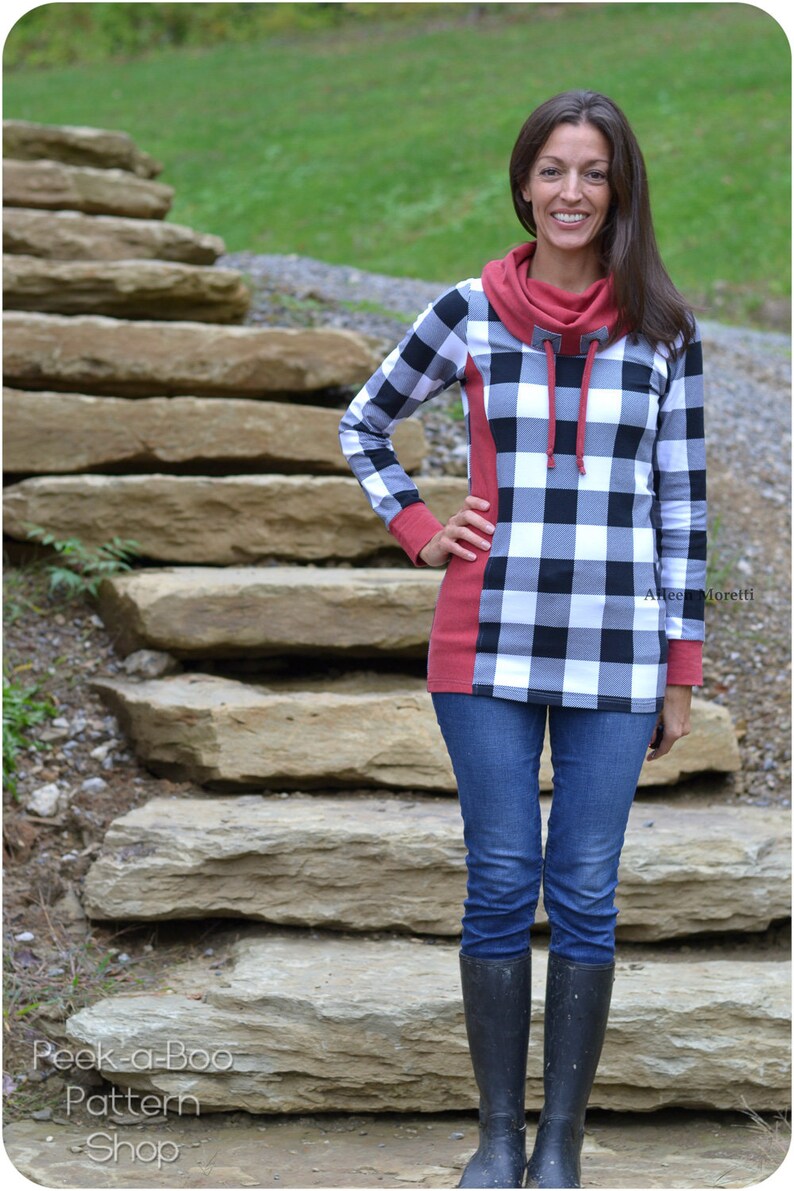 May include: A woman wearing a black and white plaid shirt with a red cowl neck and long sleeves. She is wearing blue jeans and black boots. She is standing in front of a stone staircase.