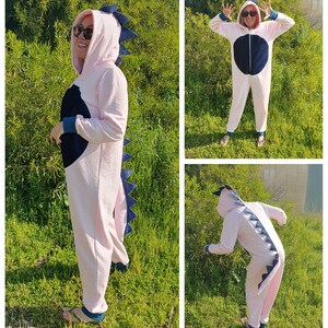 May include: Pink dinosaur onesie with a blue tail and a black circle on the chest. The onesie has a hood with a blue dinosaur head.