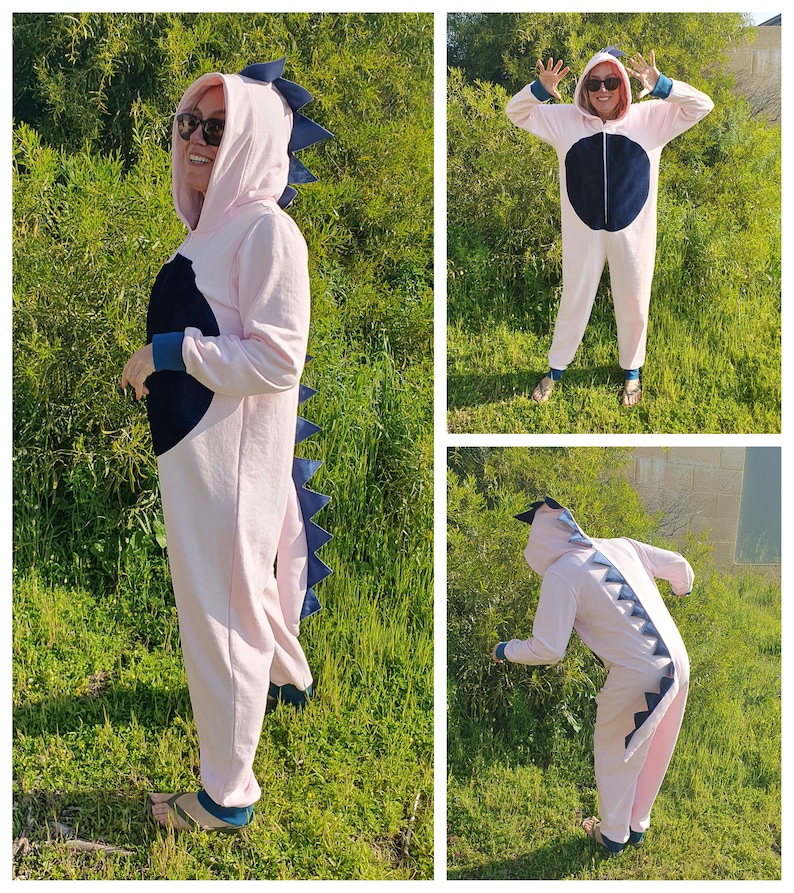 May include: Pink dinosaur onesie with a blue tail and a black circle on the chest. The onesie has a hood with a blue dinosaur head.