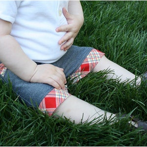 May include: A child wearing a white shirt and denim shorts with a red and white plaid trim. The child is sitting in a grassy area.