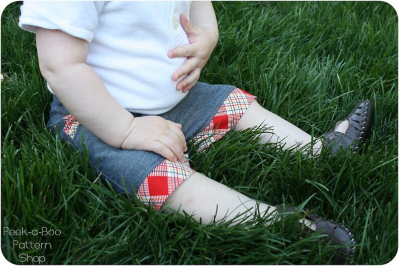 May include: A child wearing a white shirt and denim shorts with a red and white plaid trim. The child is sitting in a grassy area.