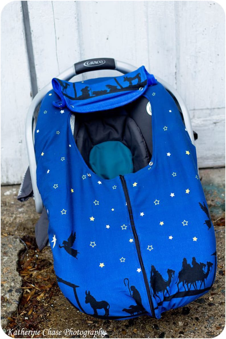 May include: A blue car seat cover with a star pattern and a nativity scene design. The cover has a zipper down the front and a flap that can be folded down.