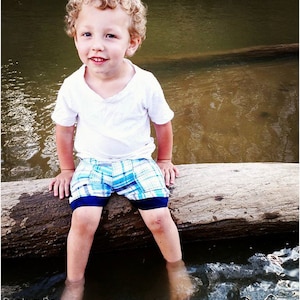 May include: A young boy with curly blonde hair sits on a log in a shallow stream. He is wearing a white t-shirt and blue and white plaid shorts. The water is clear and the log is brown and weathered.