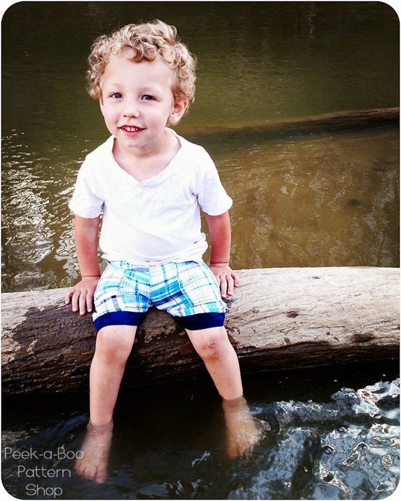 May include: A young boy with curly blonde hair sits on a log in a shallow stream. He is wearing a white t-shirt and blue and white plaid shorts. The water is clear and the log is brown and weathered.