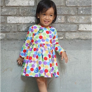 May include: A young girl wearing a white dress with colorful polka dots and cartoon faces. The dress has long sleeves and a gathered skirt. She is standing in front of a brick wall.