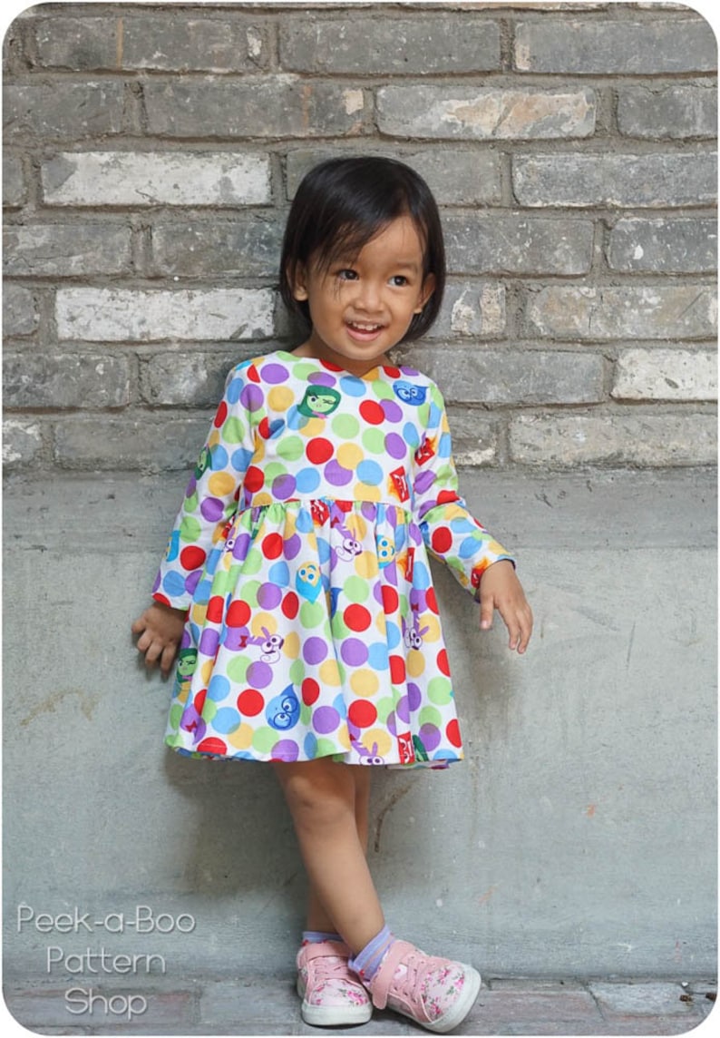 May include: A young girl wearing a white dress with colorful polka dots and cartoon faces. The dress has long sleeves and a gathered skirt. She is standing in front of a brick wall.