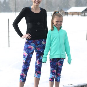 May include: Two people wearing matching athletic wear. One is wearing a black long-sleeve shirt and galaxy print capri leggings. The other is wearing a mint green zip-up jacket and galaxy print capri leggings. The background is a snowy landscape.