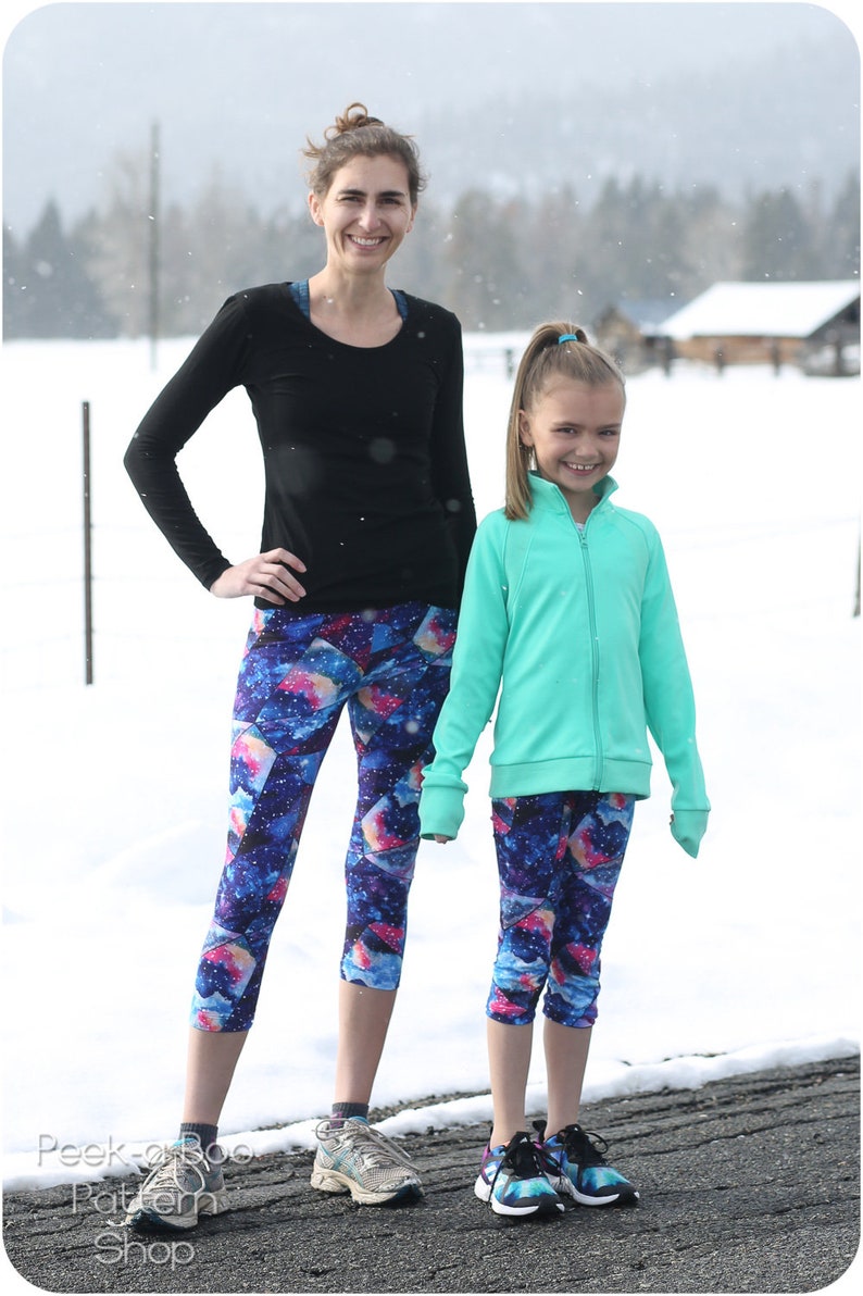 May include: Two people wearing matching athletic wear. One is wearing a black long-sleeve shirt and galaxy print capri leggings. The other is wearing a mint green zip-up jacket and galaxy print capri leggings. The background is a snowy landscape.