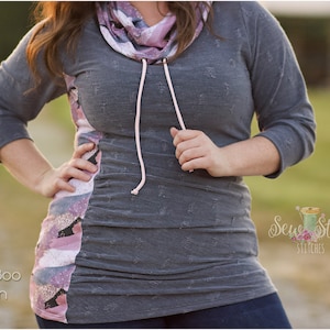 May include: A gray and pink long-sleeved shirt with a cowl neck and a drawstring. The shirt has a distressed look and is made of a soft, comfortable fabric. The shirt is shown on a model in a field.