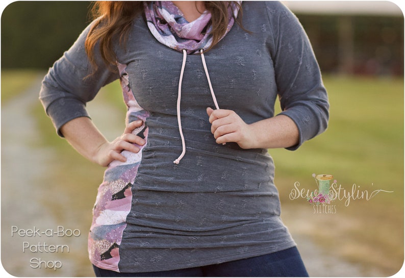 May include: A gray and pink long-sleeved shirt with a cowl neck and a drawstring. The shirt has a distressed look and is made of a soft, comfortable fabric. The shirt is shown on a model in a field.