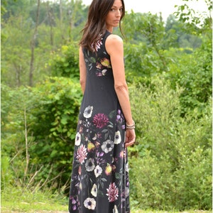 May include: A woman wearing a sleeveless maxi dress with a floral print. The dress is black with a variety of colorful flowers, including pink, purple, white, and yellow. The dress has a fitted bodice and a flowing skirt.