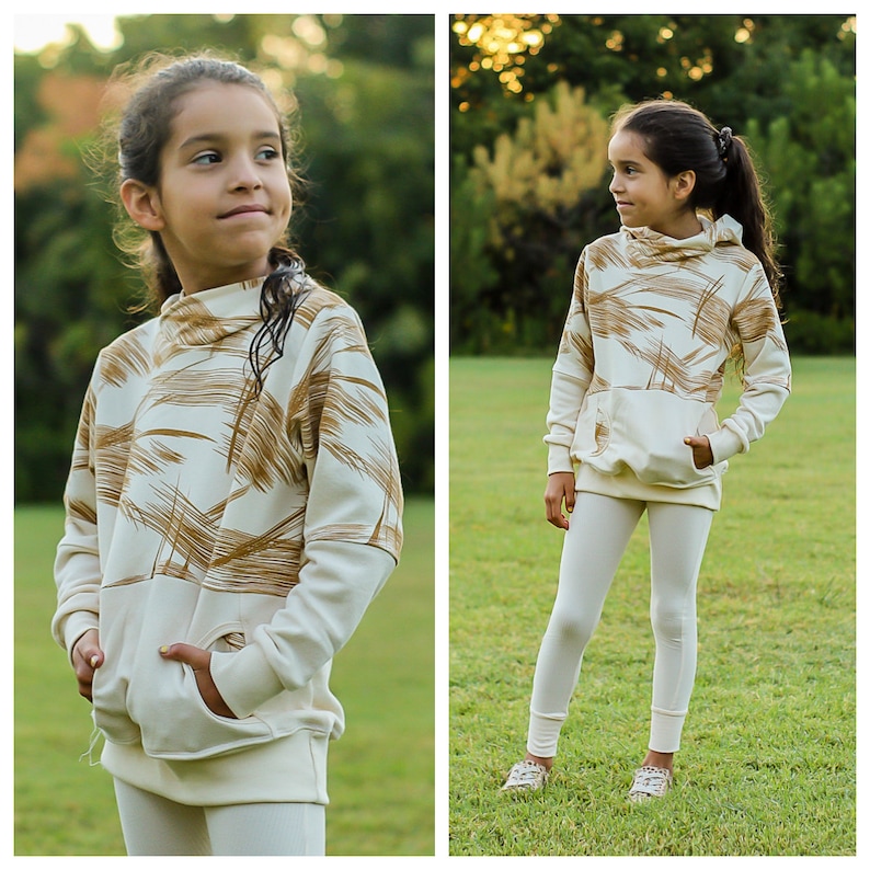 May include: A young person wearing a white and brown patterned hoodie with a white turtleneck. The hoodie has a kangaroo pocket and long sleeves. The person is also wearing white leggings.