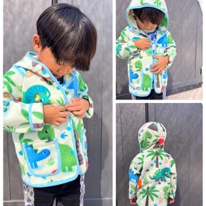 May include: A light green fleece jacket with a blue trim and a dinosaur print. The jacket has a hood and a front zip closure.