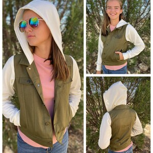 May include: An olive green hooded vest with white sleeves and a white hood. The vest has a button-up front and two chest pockets. The person is wearing a pink shirt and blue jeans. The person is wearing sunglasses.