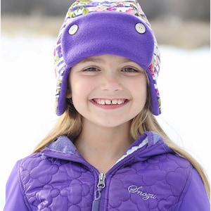 May include: A young girl wearing a purple fleece-lined hat with a floral print and a purple quilted jacket with the word "Snozu" embroidered on the front.