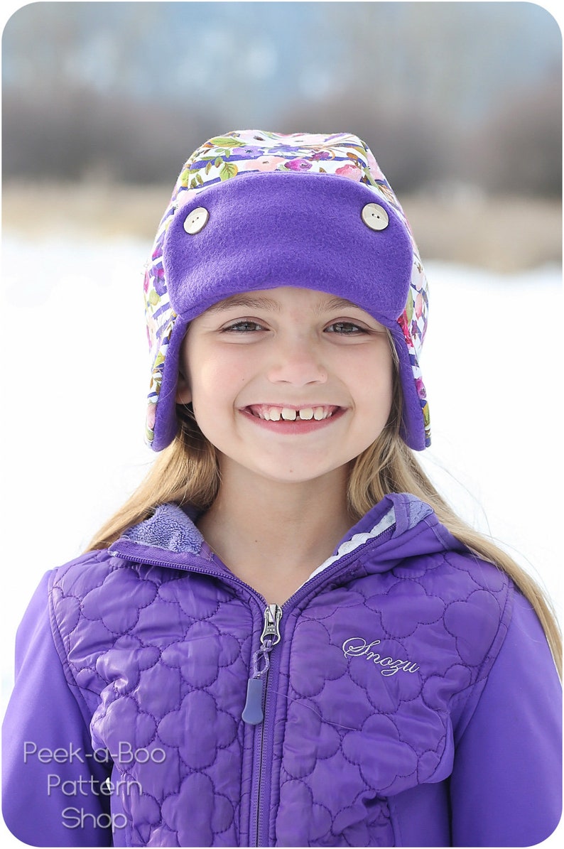 May include: A young girl wearing a purple fleece-lined hat with a floral print and a purple quilted jacket with the word "Snozu" embroidered on the front.