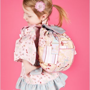 May include: A young girl wearing a pink and white floral dress with a ruffled hem, holding a patchwork backpack with a pink and white floral design. The backpack has a drawstring closure and a flap with a buckle.
