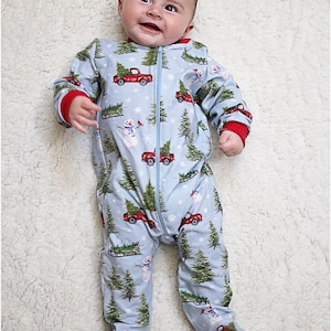 May include: A baby wearing a blue and red Christmas pajamas with a pattern of red trucks carrying Christmas trees, snowmen, and snowflakes. The pajamas have a zipper down the front.