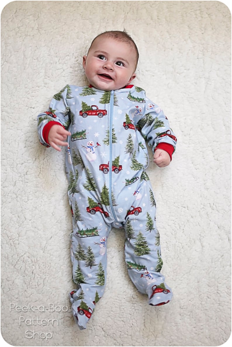 May include: A baby wearing a blue and red Christmas pajamas with a pattern of red trucks carrying Christmas trees, snowmen, and snowflakes. The pajamas have a zipper down the front.