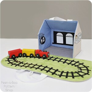 Peut inclure: Un train jouet avec une locomotive et des wagons en bois sur un tapis de jeu vert avec des rails de train noirs. Le train jouet est devant une gare en tissu bleu et blanc avec les mots "Tickets" et "Baker City" imprim&eacute;s dessus.